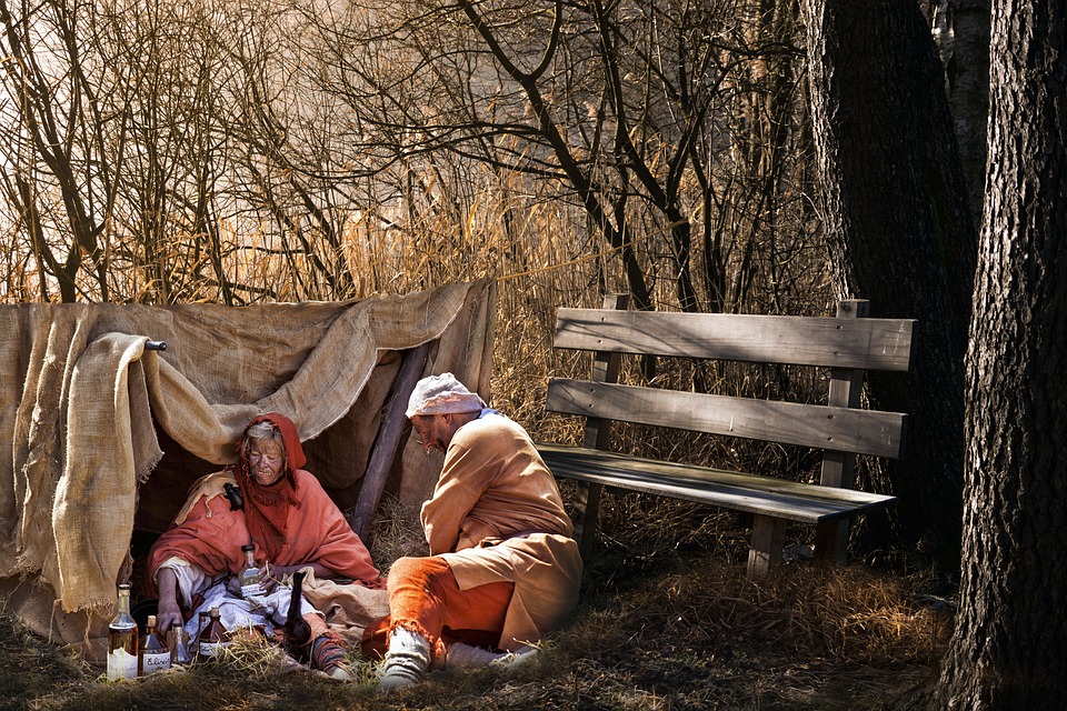 NLP Two people in worn clothing sit by a makeshift shelter of burlap, surrounded by trees. Bottles lie nearby, and an empty bench sits to their right. The scene evokes a sense of healing and recovery in a quiet wooded setting. Hypnotherapy